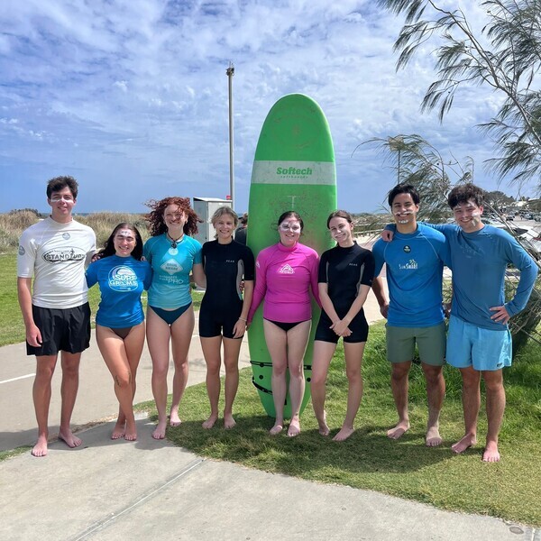 A group of young adults pose with a surfboard on the beach, wearing swimwear and with white markings on their faces.